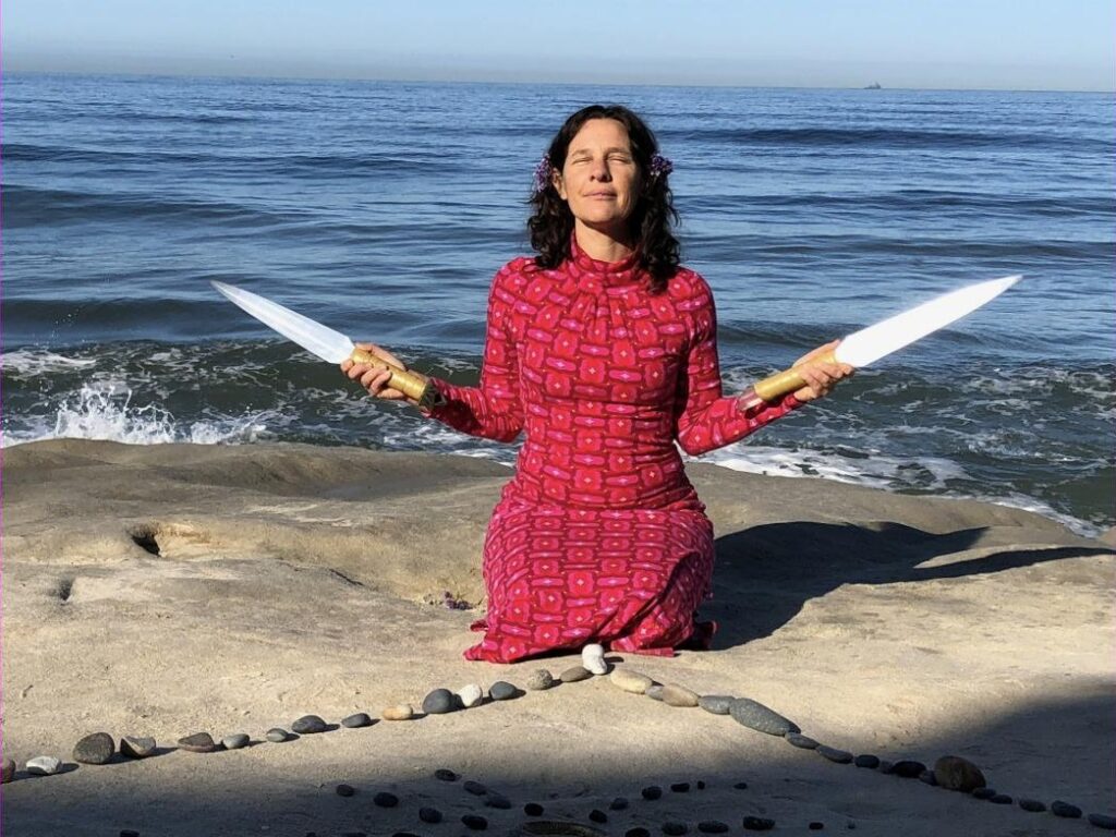A practitioner kneeling on a coastal rock platform holding a Selenite Sword of Light in each outstretched hand