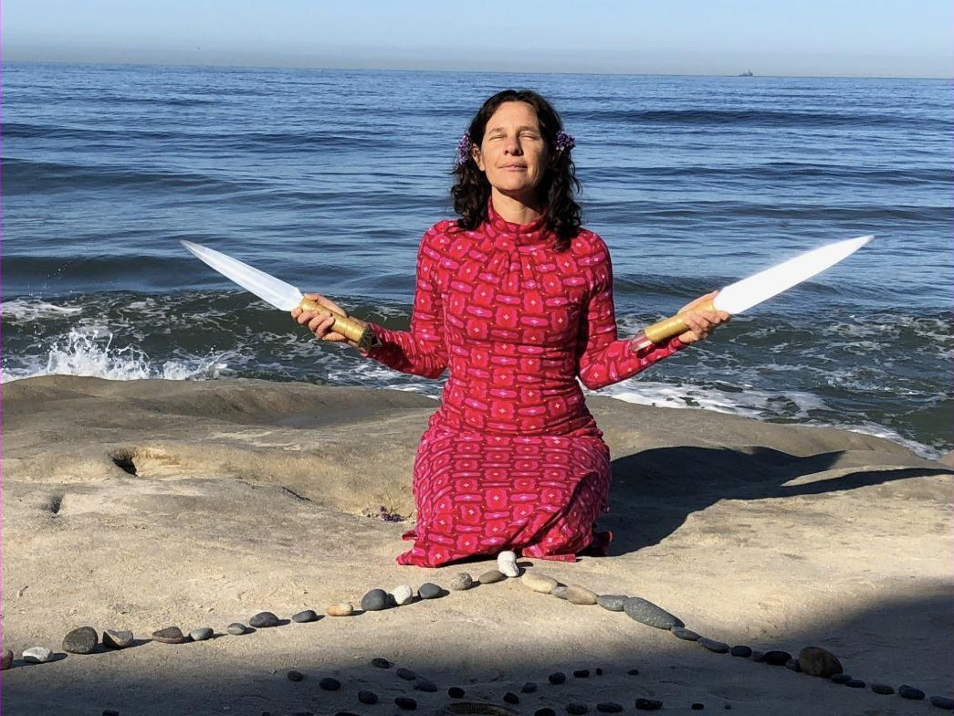A practitioner kneeling on a coastal rock platform beside a stone spiral, holding a Selenite Sword of Light in each outstretched hand with the ocean behind