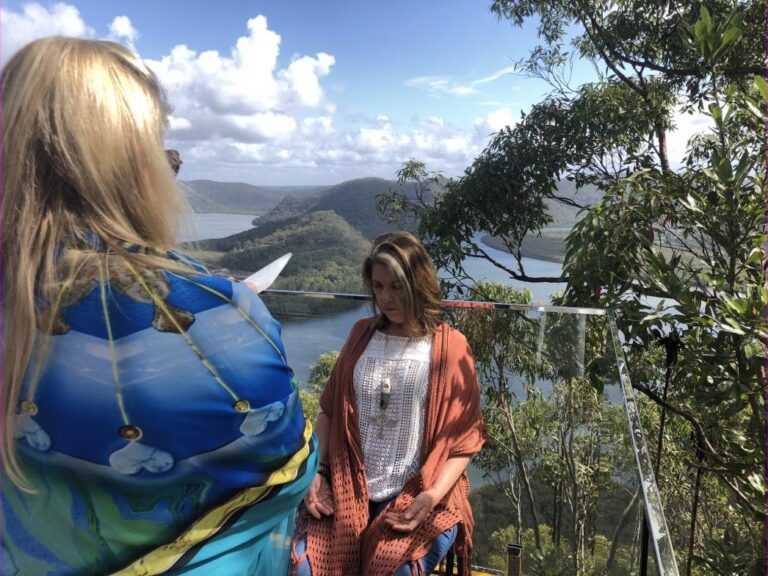 Two practitioners on a timber lookout above a wide river valley of forested hills, one holding a Selenite Sword of Light