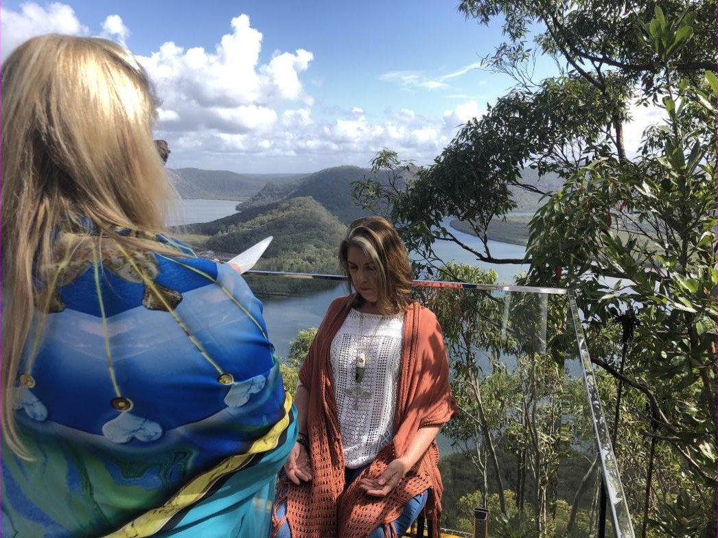 Two practitioners on a timber lookout above a wide river valley of forested hills, one holding a Selenite Sword of Light