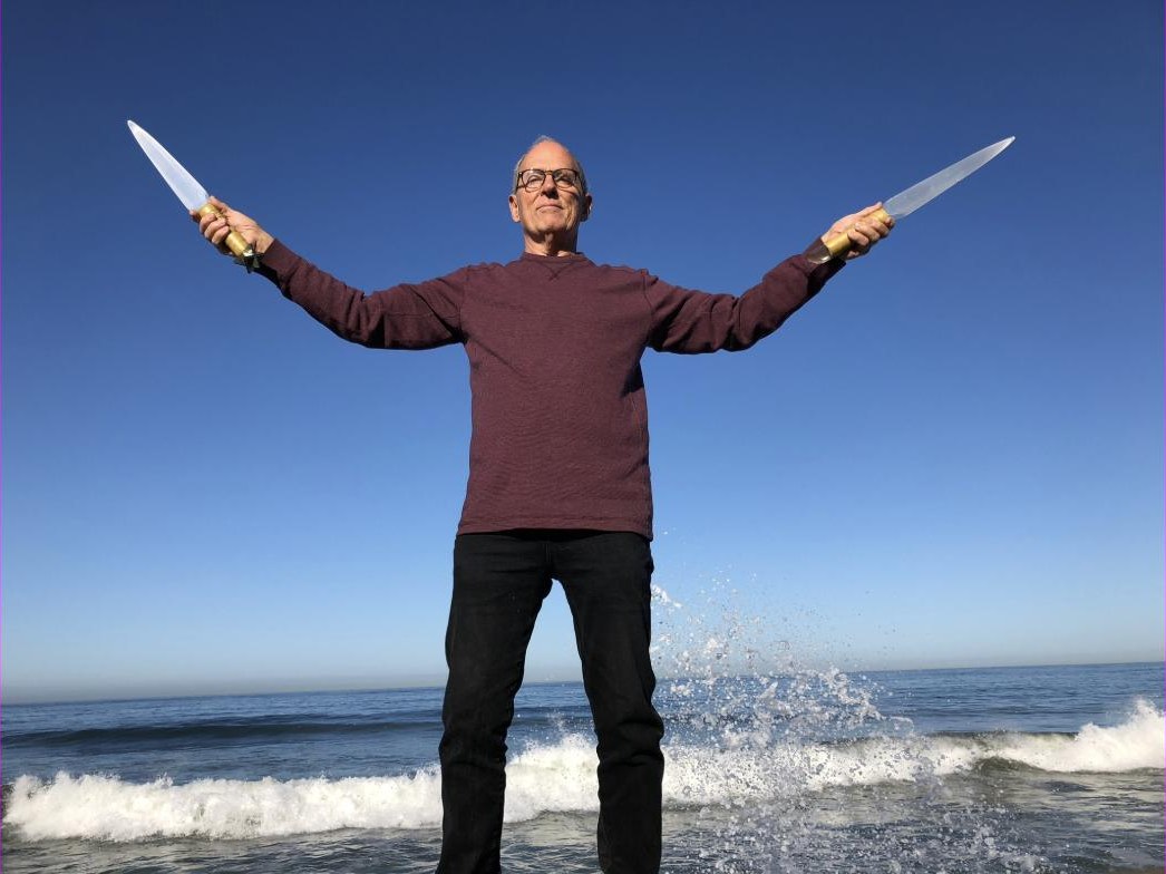 Tom Ledder standing at the shoreline with arms outstretched, holding a Selenite Sword of Light in each hand as a wave breaks behind him